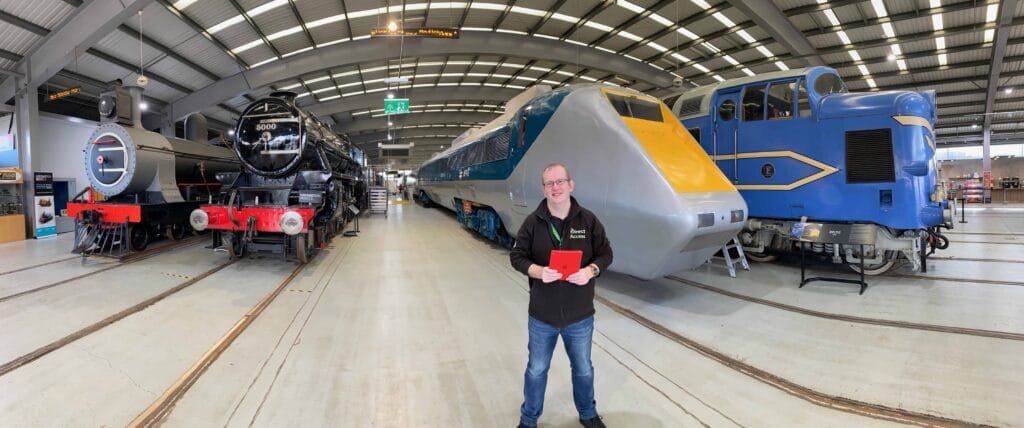 A smiling Caucasian man, Steve Dering, holding an iPad is stood in front of various old trains at the Science and Industry Museum.
