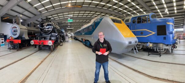 A smiling Caucasian man, Steve Dering, holding an iPad is stood in front of various old trains at the Science and Industry Museum.