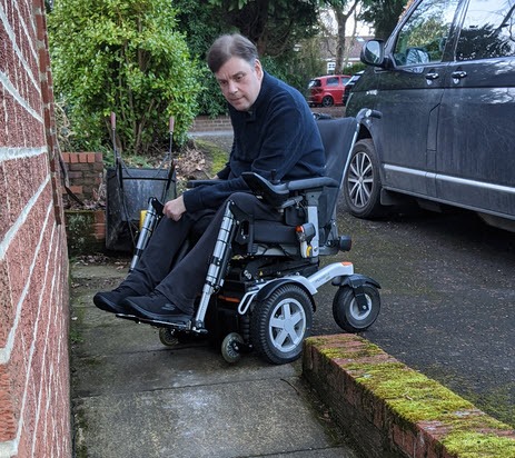 A photograph of Senior Access Consultant Tom Morgan inspecting a ramp on-site at a public facility. Tom is wearing a blue coat and is in a wheelchair.