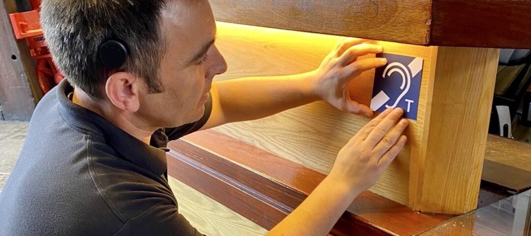 A man places a Hearing Loop sticker onto the underside of a desk.