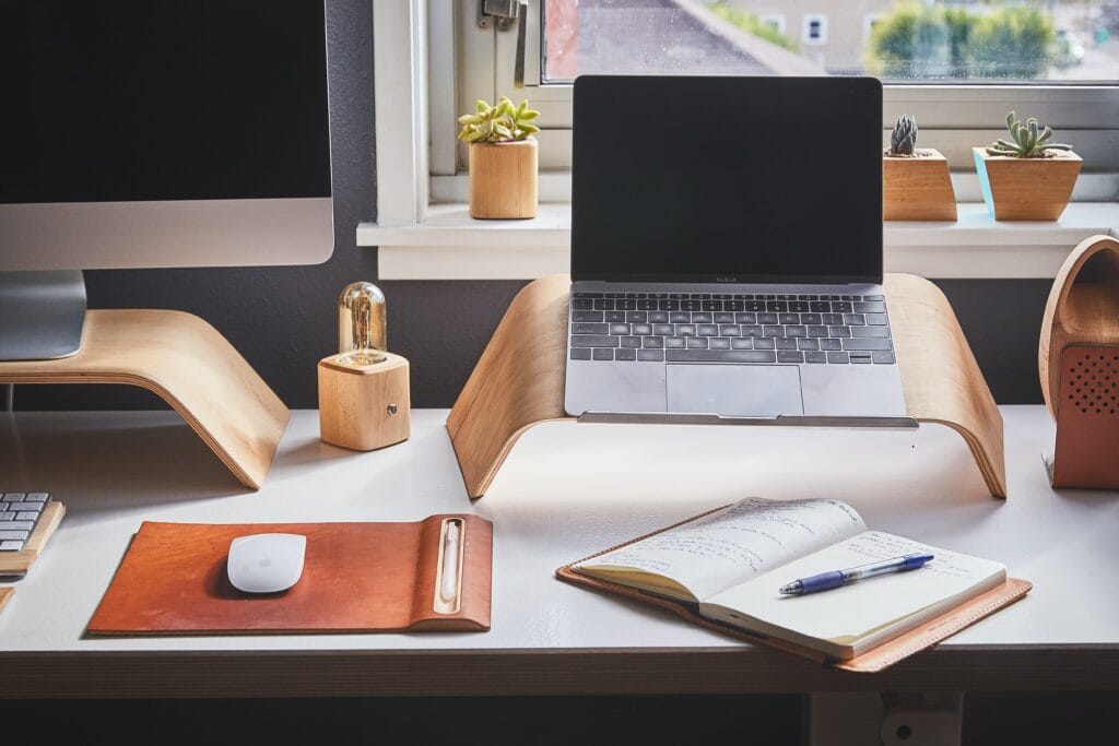 A person's workstation featuring a Mac computer, small laptop on a wooden stand, and an open notepad with a ballpoint pen sat atop it. In the background, a windowsill with a modest collection of cactus plants.