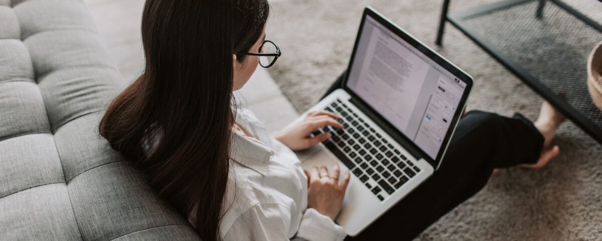 A young woman in her living room is sitting on the floor leaning against a grey sofa while doing coursework on a laptop.