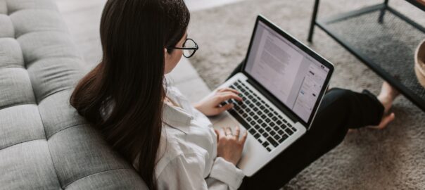 A young woman in her living room is sitting on the floor leaning against a grey sofa while doing coursework on a laptop.