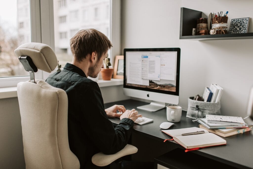 A young white man with short brown hair sits at a desk typing on his Mac computer. On his desk are numerous notepads and a cup of coffee.