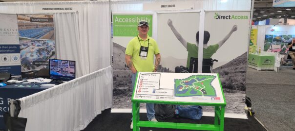 A middle aged man, Andy Lomas, poses for a photo behind a large tactile map board at a product and service conference. In the background is a poster banner for Direct Access showing a young man in a wheelchair cheering on a hillside.