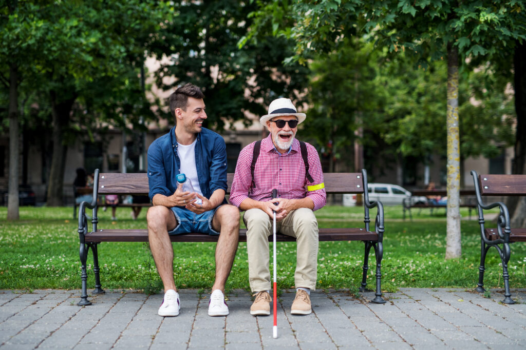 A young man and a blind senior citizen holding a white cane sit on bench in park in city.