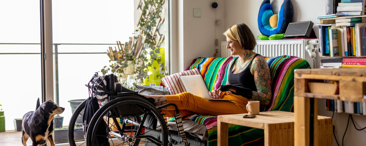A smiling young woman with a tattoo sleeve on her living room sofa typing on a laptop with her feet held up on a wheelchair. Her brown and black dog watches her.