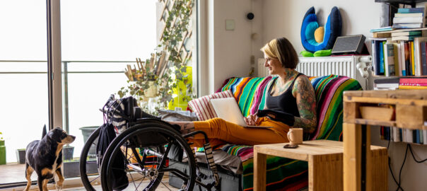 A smiling young woman with a tattoo sleeve on her living room sofa typing on a laptop with her feet held up on a wheelchair. Her brown and black dog watches her.