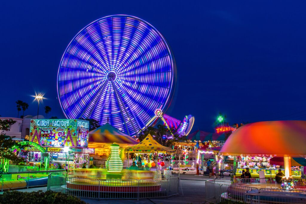 A time-lapsed photograph of a theme park at night time, likely during final hours. The colourful rides glow in the night, the most impressive being the big wheel glowing purple as it spins.