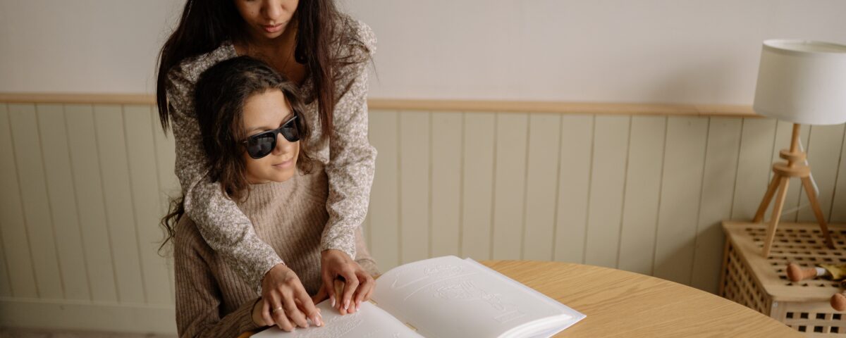 A young woman helps a female blind child interpret the image of a hedgehog in a braille book.