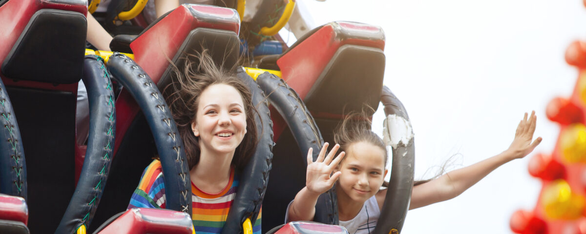 Two female children smile as a rollercoaster sends them flying down the track. One of them is posing for the camera holding her hands up high.