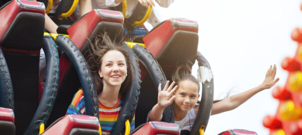 Two female children smile as a rollercoaster sends them flying down the track. One of them is posing for the camera holding her hands up high.