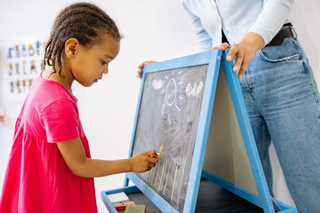 A young mixed race schoolgirl with dreadlocks drawing a bee on on a small chalkboard with a yellow piece of chalk while being supervised by a young white woman.