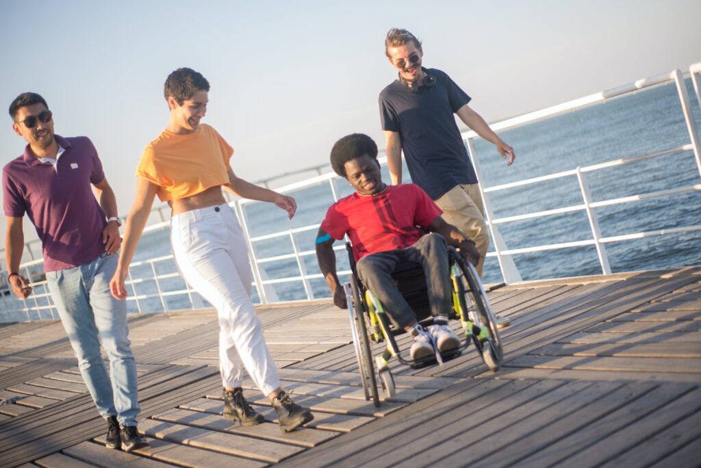 A mixed group of young people happily walk along a pier with the calm blue sea behind them. One of them, a young black man, is in a wheelchair.