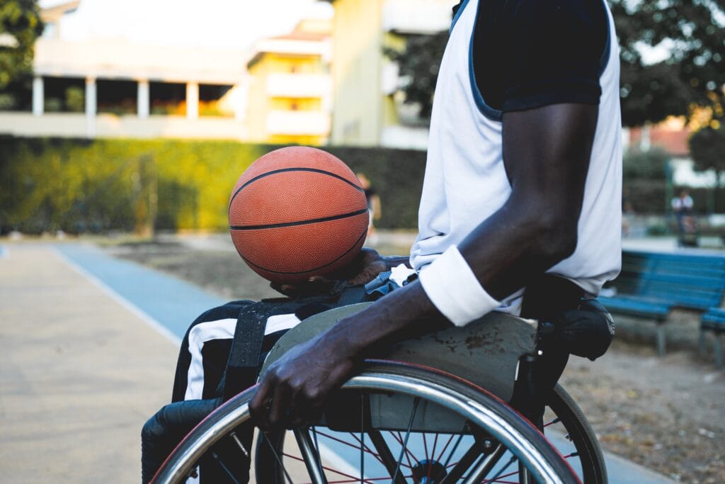 A black wheelchair user wearing a white vest and armband carries a basketball in the palm of his left hand and holds on to his wheelchair with his right. He is on a basketball court on a sunny day.