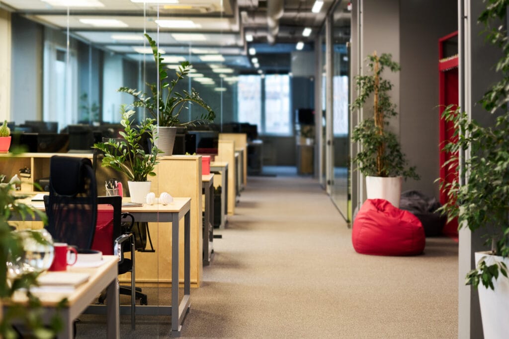 Part of a spacious office with a long aisle and row of desks with business supplies and green plants standing by walls and on workstations.