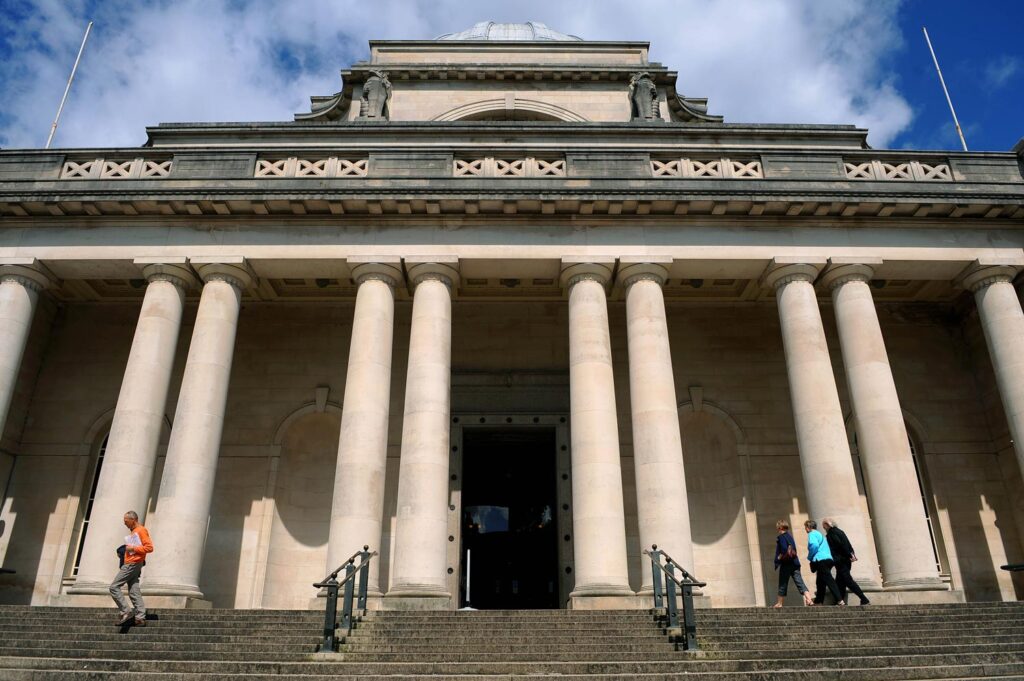 An exterior view of the front of the National Museum in Cardiff on a cloudy day. A grand Greek style building with pillars supporting the front entrance ceiling.