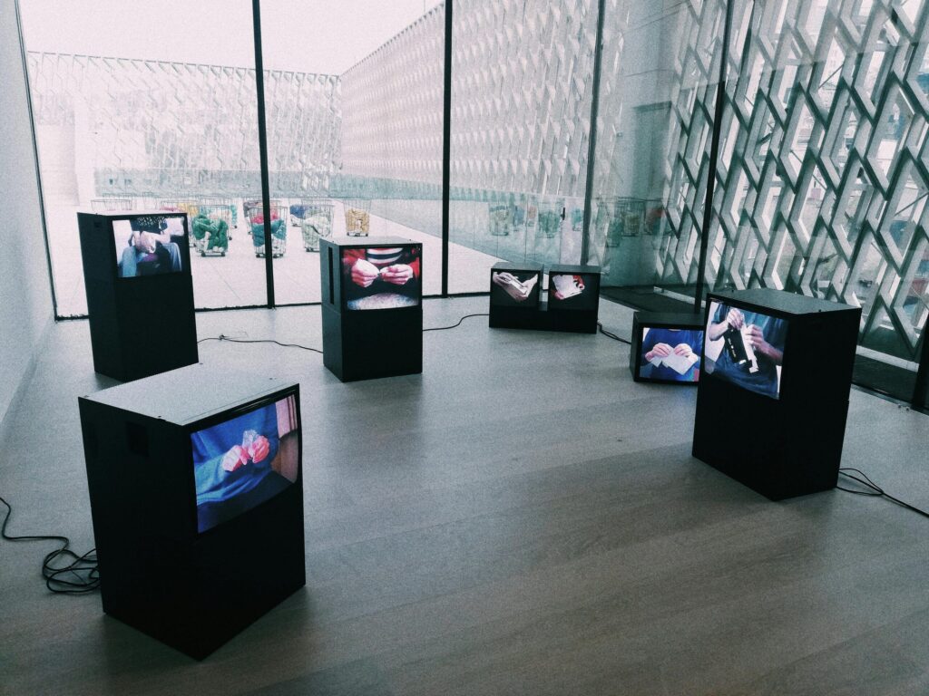A general wide view of a modern art exhibition, with several TV screens of different sizes and shapes scattered across an otherwise empty floor. On each of the TV's, people's hands are shown handling or tearing different items.