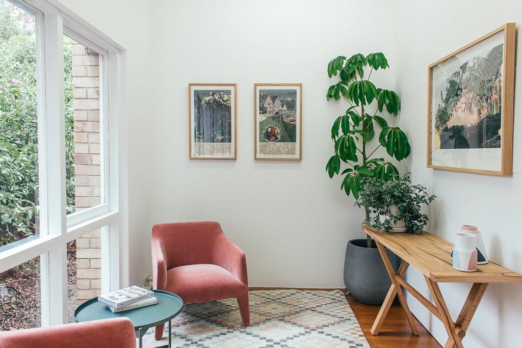 A modern living room space with artificial plants decorating a space which includes a coffee table and red armchairs.