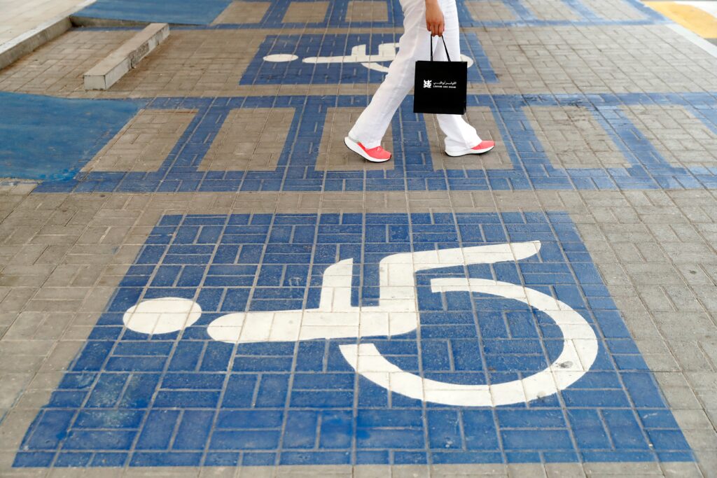 The international symbol of access (white wheelchair symbol) is painted on stone slaps on a walkway. The bottom half of a woman can be seen, she is wearing white jeans and is carrying a black bag.
