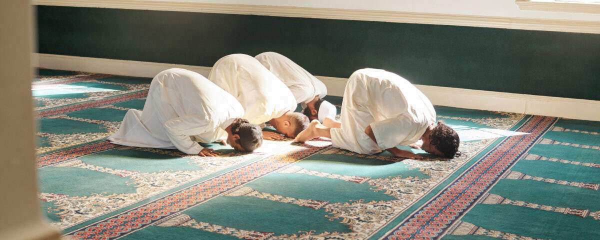 A group of Muslim bowing down during prayer in a Mosque as the sunlight shines through on to their white garments.