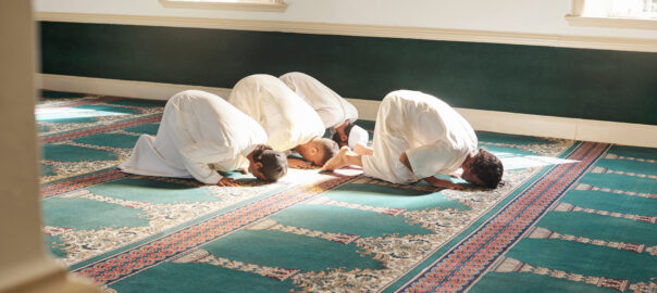 A group of Muslim bowing down during prayer in a Mosque as the sunlight shines through on to their white garments.