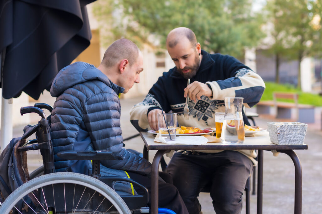 A young Caucasian man who is a wheelchair user eats on the terrace of a restaurant with a Caucasian male friend. The friend assists by cutting his food up with a knife and fork.