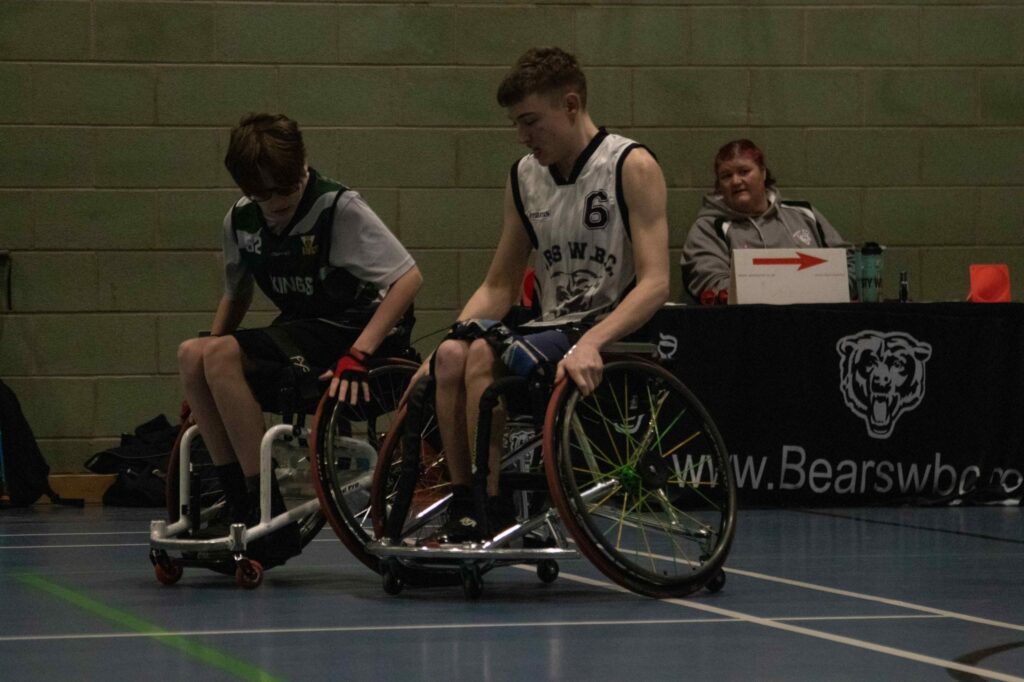 A photograph of two young male and caucasian teenage wheelchair users in dark blue and white sportswear sit on the corner of an indoor basketball court. A bench with a team captain/coach, a middle aged woman, is sat at a table behind them observing the match.