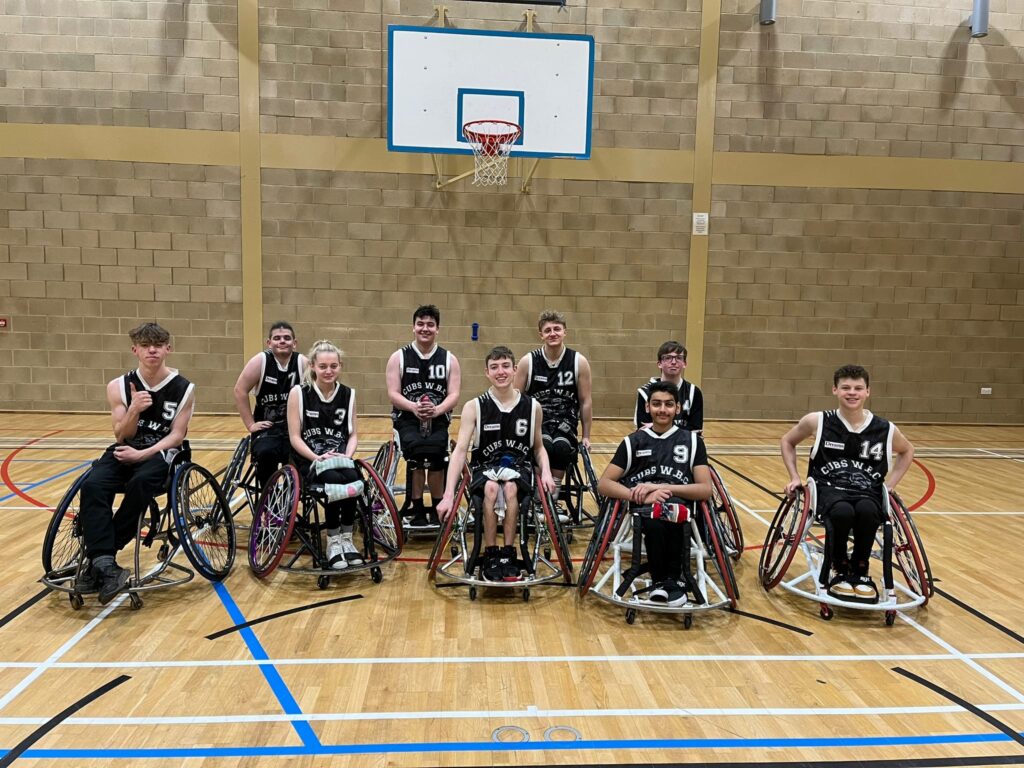 A mixed group of teenage wheelchair users within an indoor basketball court smile as they pose for a photo. Above them is a basketball hoop.