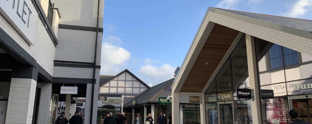A photograph of Cheshire Oaks retail outlet on a sunny afternoon. A few cloud dot the otherwise blue sky as shoppers walk along the promenade. To the right of the image are Dune and Clogau stores.
