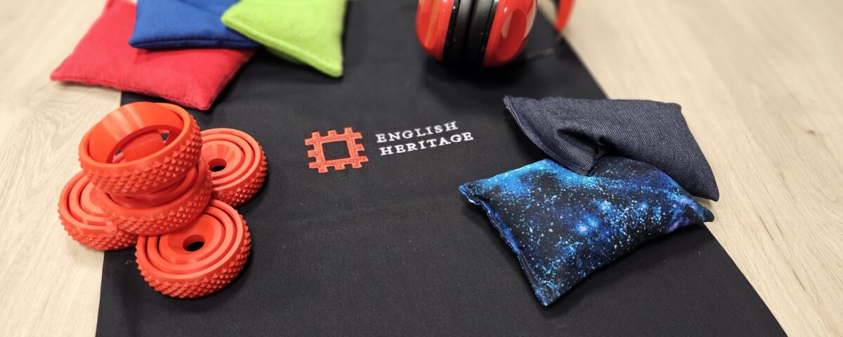 An image of a black tote bag on a table with the English Heritage logo on it. Surrounding the tote bag are various items relating to accessibility, including ear defenders, bean bags and squeezable rings.