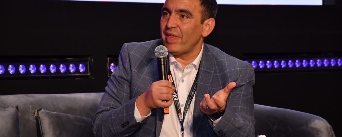 A middle aged Maltese man in a pink polo shirt and light-blue checkered suit jacket sits on a sofa in an auditorium. He is holding a microphone and holding his hand out as he says something to an off-screen interviewer.