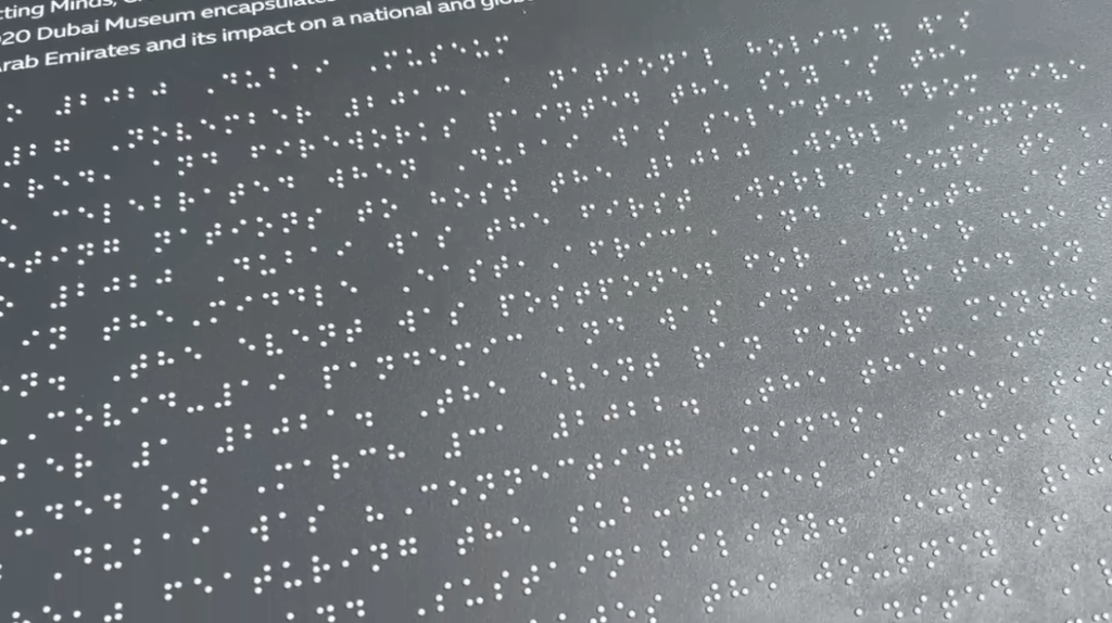 A close-up photograph of the raised braille embedded on a map as the sun shines and reflects of its otherwise grey, flat surface.