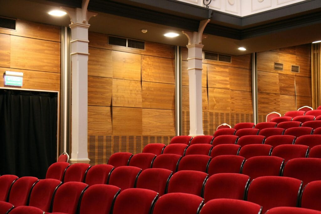 The interior of an empty movie theatre warmly lit by overhead lights. In the forefront of the photograph are empty seats all red in colour.