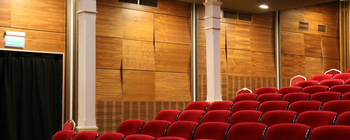 The interior of an empty movie theatre warmly lit by overhead lights. In the forefront of the photograph are empty seats all red in colour.