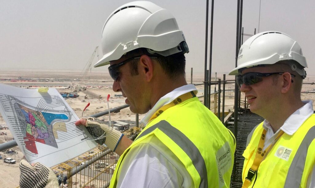 Two middle aged men overlooking a giant building site in the desert look through design drawings on a piece of paper.