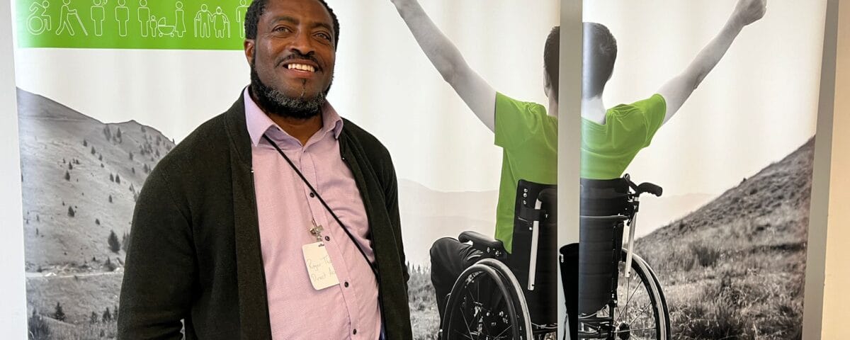 WhatsApp Image 2024-07-03 at 17.11.53 A black man wearing a pink polo shirt, grey trousers, and a black cardigan poses in front of a Direct Access exhibition banner which has a grayscale image of a young man in a wheelchair on a cliffside holding his arms in the air.
