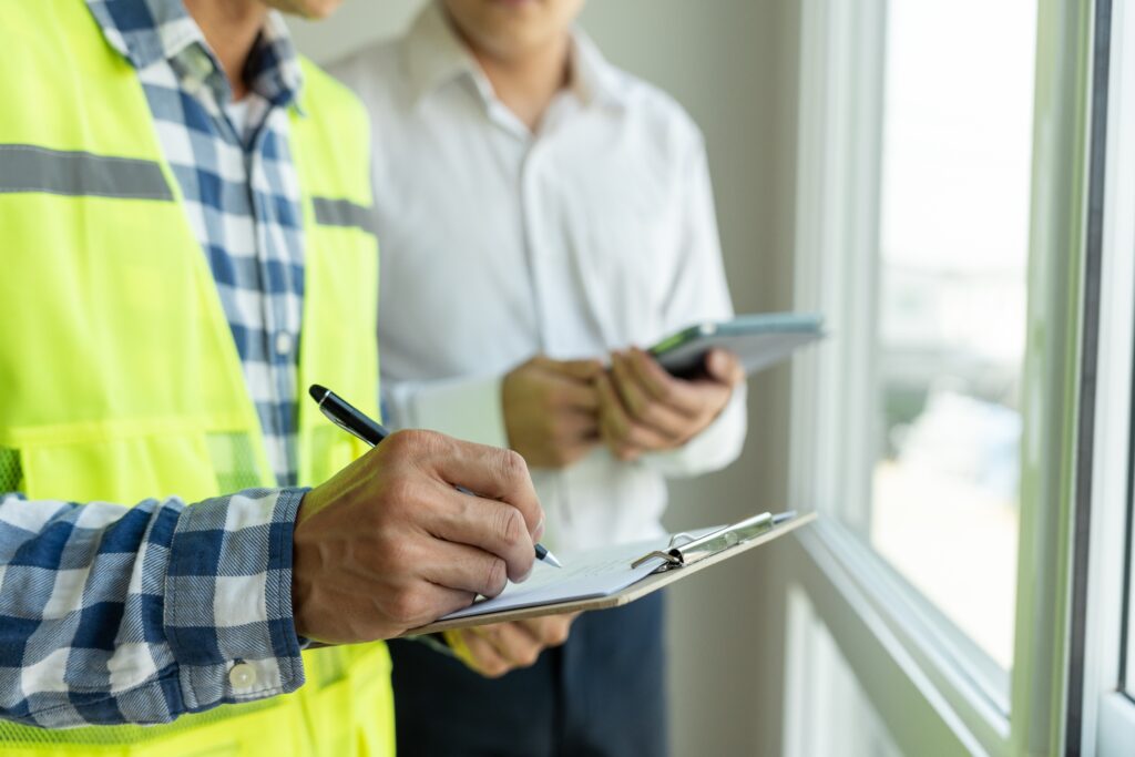 An inspector holding a pen and clipboard writes down something while an inspector holding an electronic tablet from behind observes. Both are stood by a windowsill from within a building.