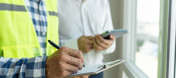 An inspector holding a pen and clipboard writes down something while an inspector holding an electronic tablet from behind observes. Both are stood by a windowsill from within a building.