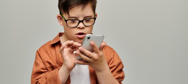 A little boy with down syndrome wearing glasses focuses intently on a mobile phone, scrolling on the screen.