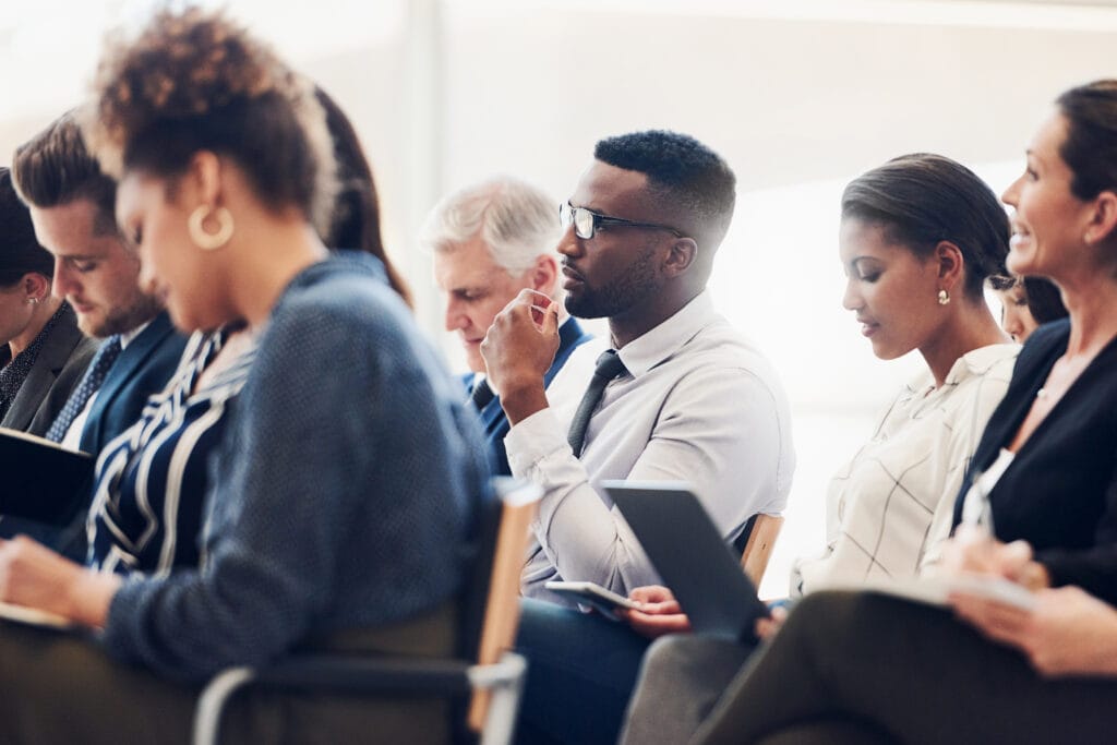 Shot of a mixed group of businesspeople sat on different rows of seats attending a class. Some are watching an out of shot person attentively, while others are writing information on their notebooks, laptops, and tablets.