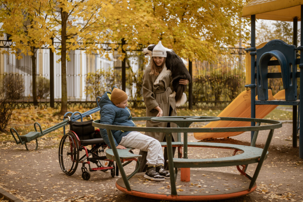 A 10-year-old boy sits on a carousel in a playground ready to be spun by his mother, who is carrying their pet dog in her other hand. Behind the pair is the boy's wheelchair. The weather is clear and the season is autumn. Brown leaves are scattered throughout the playground.