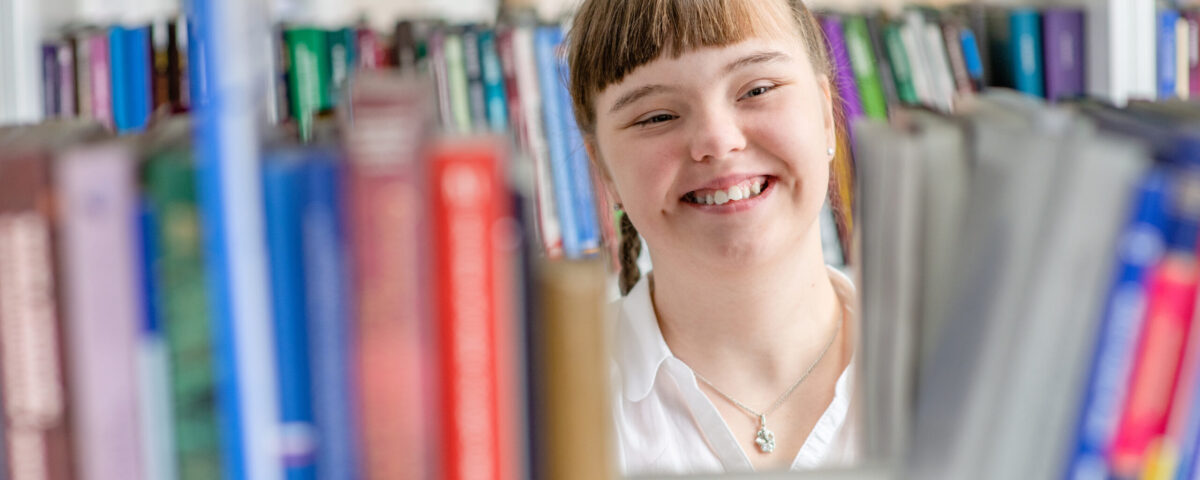 Caucasian girl with down syndrome smiles while choosing a book on a shelf in a school library.