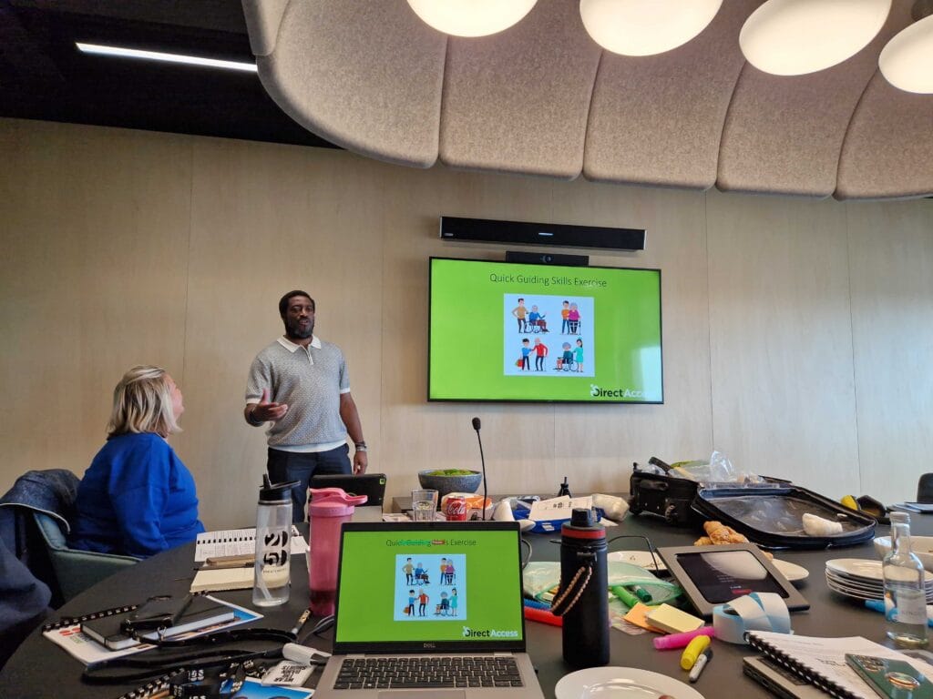 A caucasian woman in a boardroom listening attentively to Roger, a black man delivering a presentation on accessibility awareness. On the television next to him is a PowerPoint.