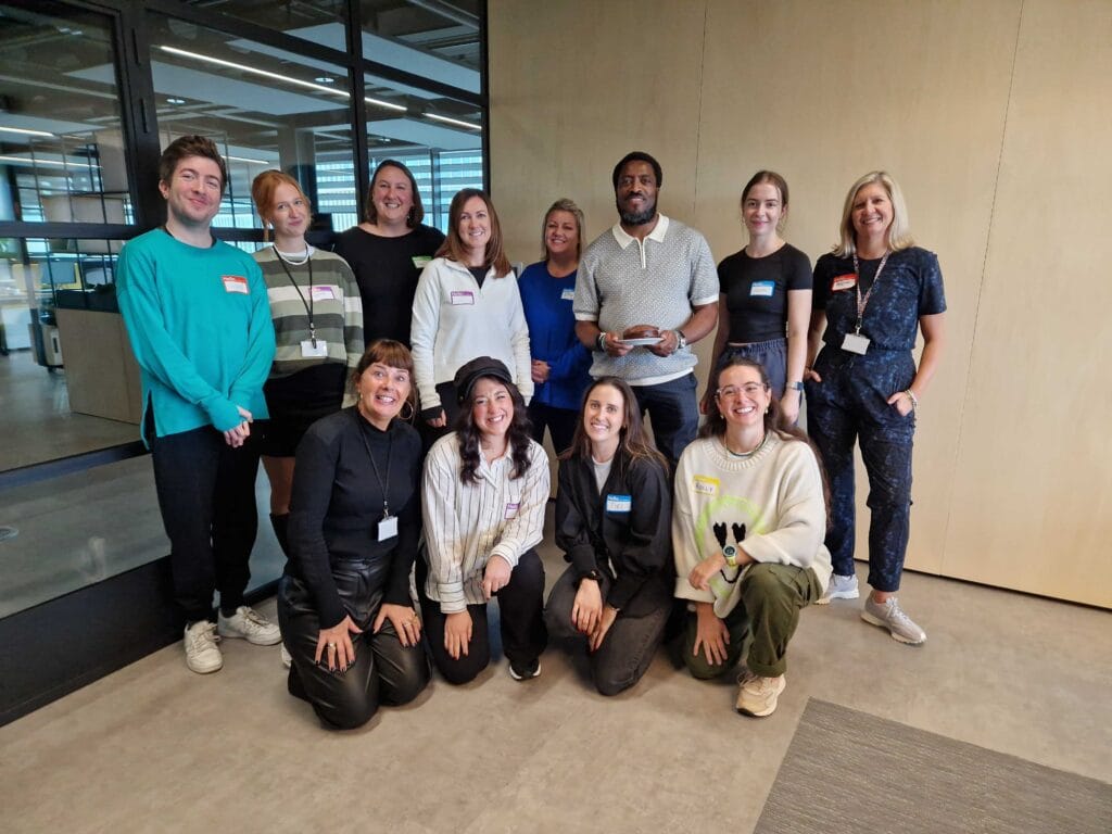 A mixed group of adults pose for a group photograph at the end of their accessibility awareness training in the corner of a boardroom. All are smiling and some people are thrusting their arms in the air energetically. A few adults are kneeling on the floor.