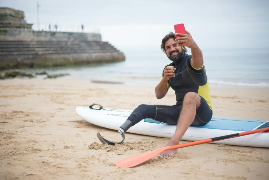 A male surfer with a mechanical leg sitting on a surfboard on a beach takes a selfie on his phone with the tide in the background.