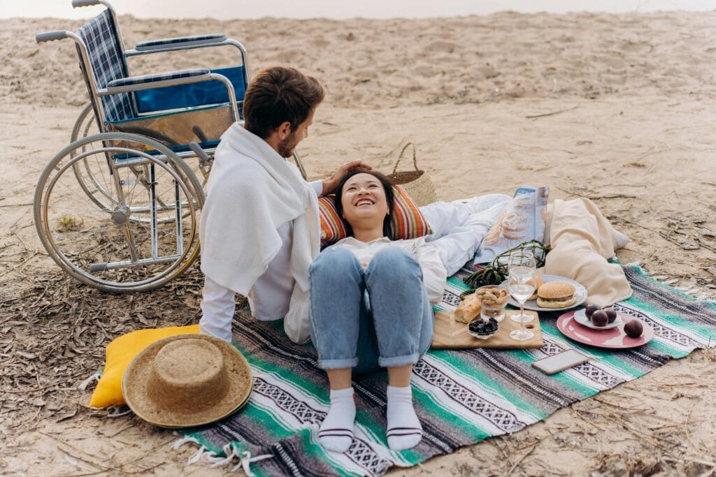 A young couple, male and female, sit on a blanket on a beach with a picnic. The woman (East Asian) is lying on her back and smiling up at her partner (Caucasian). A wheelchair is located to his left.