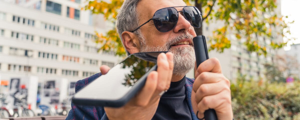 Blind middle-aged man with gray hair and beard wearing sunglasses holding cane and phone using text-to-speech function. Accessibilty. Modern technology. Horizontal outdoor shot Blind middle-aged man with gray hair and beard wearing sunglasses holding cane and phone using text-to-speech function. Horizontal outdoor shot.