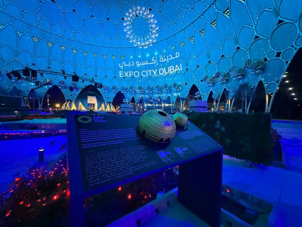 A medium shot of the interior of the Al Wasl Dome at Expo City Dubai at night time, with a Tactile Braille Map board developed by Direct Access in the center of the image. The large megastructure dome is lit up in a sea blue colour which reflects on the map board. The map board has a small tactile model of the Al Wasl dome in its center as well as text and braille.