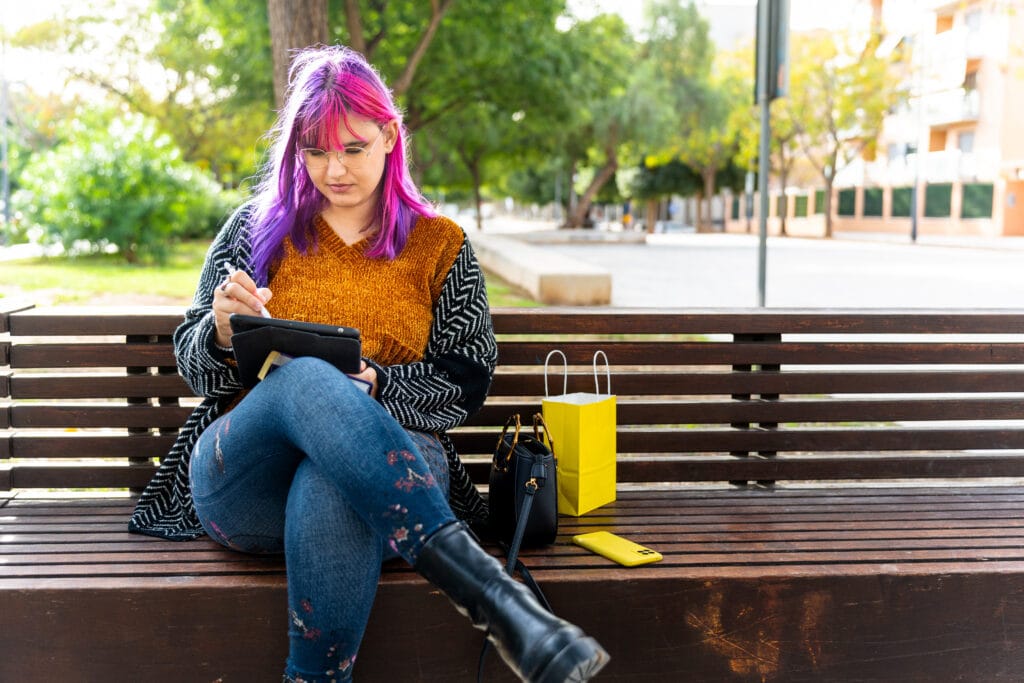 A young woman with colorful purple and pink hair uses a digital tablet and stylus on a park bench, with a shopping bag and smartphone next to her.